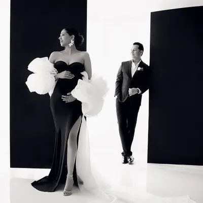 A moment of high fashion maternity. The expecting mother looks absolutely breathtaking in a black gown with dramatic white floral sleeves, creating a striking silhouette against the minimalist backdrop.