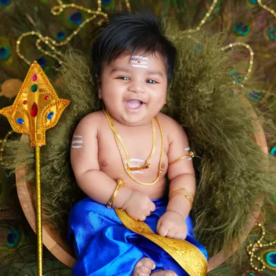 A joyful little Murugan. This photo captures the spirit of celebration with a beautiful smile, traditional attire, and props like the Vel and peacock feather background.