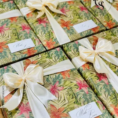 A set of gifts wrapped in a lush, tropical-print paper. The vibrant green and pink floral design is balanced with a classic cream satin bow and a simple "With Love" tag, perfect for a destination wedding or summer event.