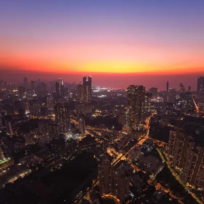 A wide-angle drone shot of the Mumbai skyline at dusk, with the city lights beginning to twinkle under a sky painted in shades of orange and purple.