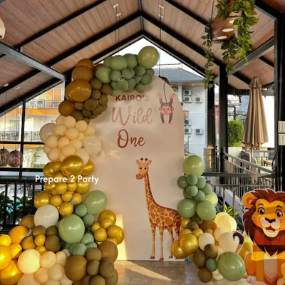 A "Wild One" jungle safari theme, perfect for a first birthday. This setup features an arched backdrop with a giraffe, a lush organic balloon garland in earthy tones, and a cute lion cutout.