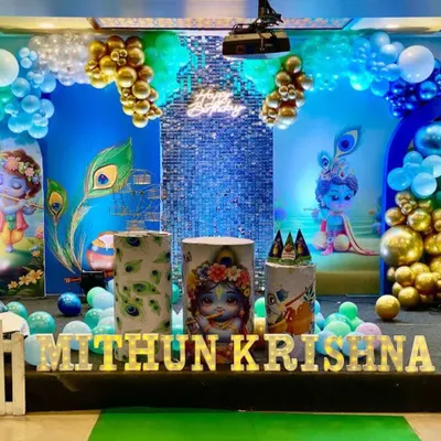 A divine Krishna-themed birthday decoration. The stage features a blue backdrop with peacock feather motifs, character cutouts of little Krishna, and a blue and gold balloon garland.