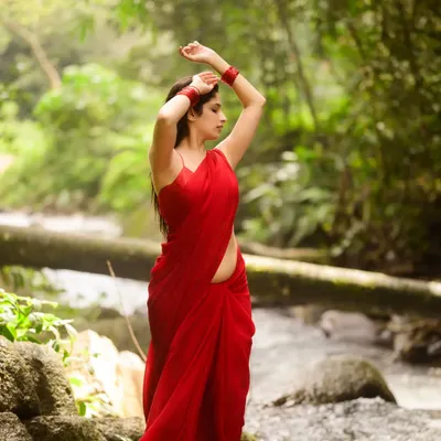 A stunning portrait in a vibrant red saree, set against the lush greenery of a forest stream. The contrast between the red and green is visually striking, creating a powerful, elemental image.