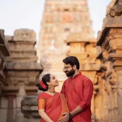 An expecting couple dressed in traditional red attire, sharing a loving gaze against the magnificent backdrop of a South Indian temple.