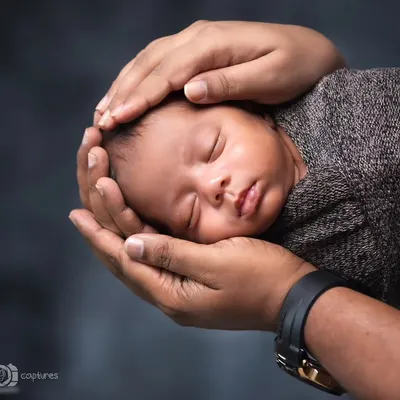 A perfect example of a safe and classic newborn pose. The baby is held securely in their parent's hands, swaddled in a neutral wrap against a dark background.
