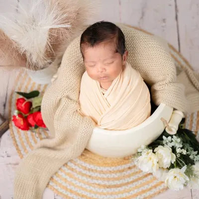 A classic and peaceful newborn portrait. This little one is perfectly swaddled and sleeping soundly in a bowl prop, surrounded by soft textures and delicate flowers for a timeless look.