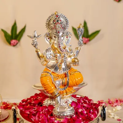This silver-plated Ganesha idol, adorned with colorful enamel details, sits amidst a bed of fresh roses. The craftsmanship highlights his divine attributes, including the conch and mace, symbolizing victory and blessings.