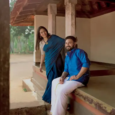 A playful, candid moment on the porch of a traditional South Indian house. The woman's blue saree and the man's mundu create a beautiful, authentic look.