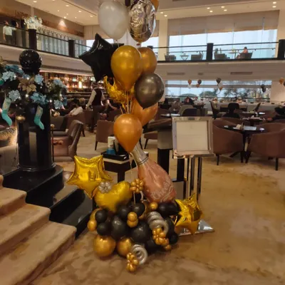 A fun and celebratory balloon bouquet perfect for congratulatory events or milestone parties. This arrangement, placed in a hotel lobby, features a large champagne bottle foil balloon, gold stars, and a base of black and gold balloons.