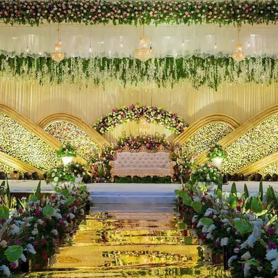 An opulent reception stage featuring golden arched panels filled with dense white flowers. The cascading floral arrangements and a grand central sofa create a look of pure elegance and luxury for a premium wedding event.