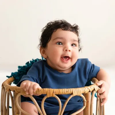 A baby with beautiful curly hair smiles brightly while sitting in a rustic wicker basket, a perfect example of a joyful sitter session.