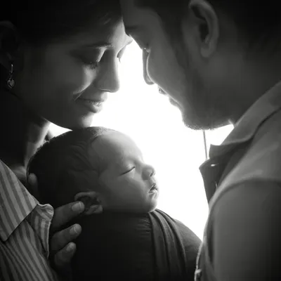 A powerful black and white portrait capturing the complete family unit. The parents gaze lovingly at their newborn, creating a silhouette effect that emphasizes their deep connection and the beginning of their new life together.