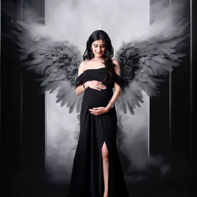 A dramatic and powerful portrait featuring a mom-to-be in a black gown with artistic angel wings, set against a moody, smoky backdrop.