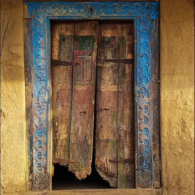 A weathered wooden door framed by an ornate, hand-carved blue border stands out against a yellow wall. The decay at the bottom hints at the countless seasons this beautiful entrance has endured.
