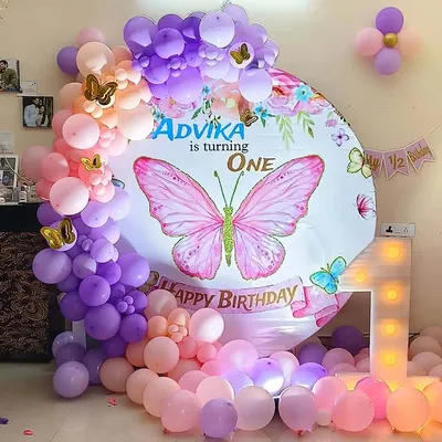 A beautiful butterfly themed first birthday decoration. The setup features a circular backdrop with a large pink butterfly, a marquee number '1', and a balloon garland in shades of pink, purple, and peach.