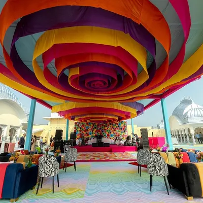 A whirlwind of color at a Carnival-themed pre-wedding event. The ceiling is a spiral of vibrant drapes, leading to a stage with a backdrop of colorful paper fans, creating an atmosphere of pure joy and celebration.