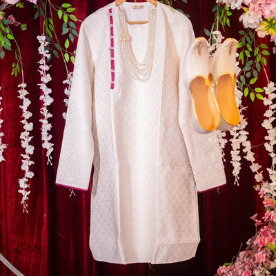 An artistic flat-lay of the groom's wedding attire, including his white kurta, pearl necklace, and traditional shoes (mojari), beautifully arranged.