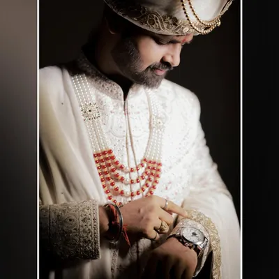 A close-up, sophisticated portrait of the groom checking his watch. The dramatic lighting highlights the rich details of his white sherwani and the intensity of the moment.