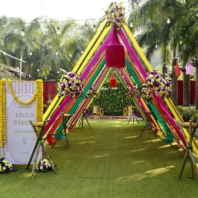 A perfect entrance for a vibrant Haldi ceremony. The welcome sign for Riya and Ishaan stands amidst a tunnel of colorful ribbons and marigolds, setting a festive tone right from the start.