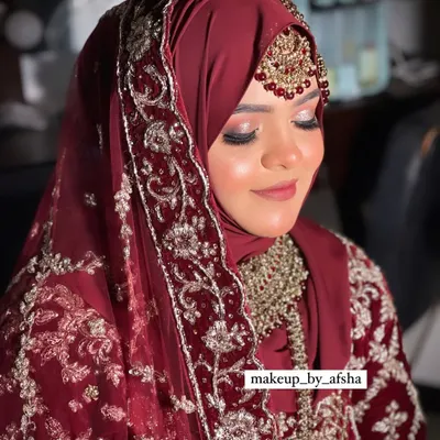 A beautiful Hijabi bride in a deep maroon outfit. The hijab is styled to perfection, and the makeup features a soft, glittery eye and a matching lip color. Mashallah, she looked so elegant.