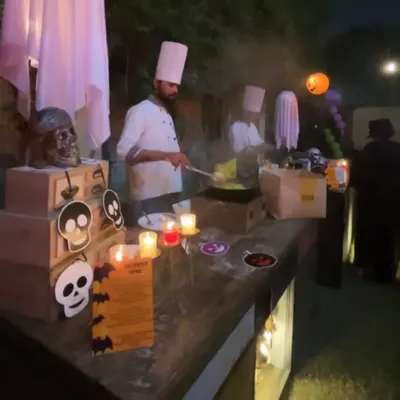 A chef at a live cooking station during a Halloween party. The station is decorated with spooky skulls, skeletons, and candles, fully immersing the cooking experience into the theme.