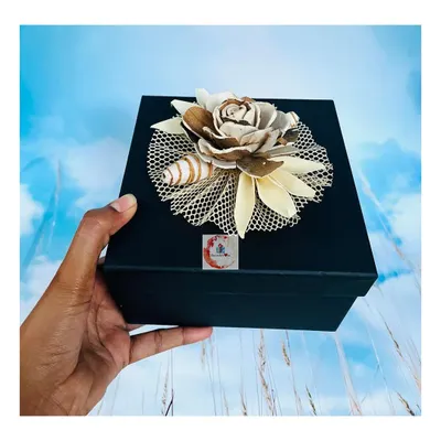 A sophisticated square black gift box, held to show its size and quality, topped with an intricate, handcrafted floral decoration made from natural materials. This is a great option for an elegant and premium feel.