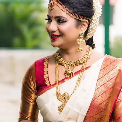 A classic South Indian bridal portrait. This bride is wearing a traditional off-white and red silk saree, complemented by antique gold temple jewellery. Her makeup features a sharp winged liner, a red bindi, and a bold red lip.