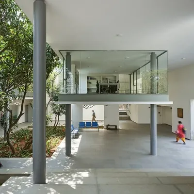 A floating glass box containing a discussion room overlooks a landscaped court at the Karunashraya Institute for Palliative Care (KIPCER). The design creates transparent, light-filled spaces for learning and collaboration.