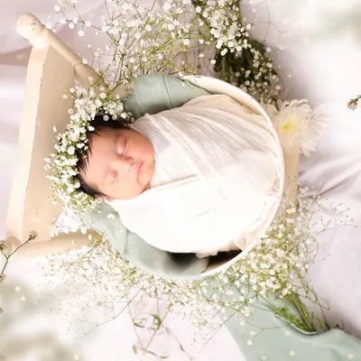 Wrapped snugly in white fabric and adorned with a floral crown of baby's breath, a newborn sleeps peacefully. This image highlights the delicate details and angelic calm of a professionally styled newborn session.
