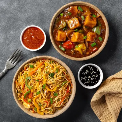 A combo of chili paneer and vegetable hakka noodles for Chhappan Bhog. The top-down angle on a dark, textured surface highlights the pairing.