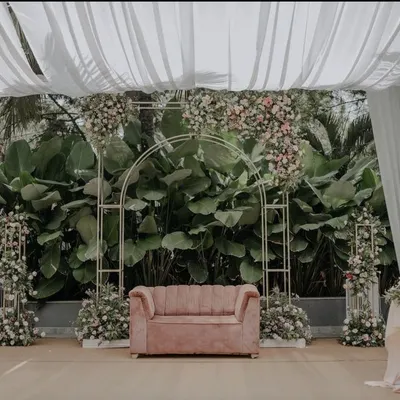 This minimalist and modern wedding stage is perfect for a chic garden ceremony. A simple gold geometric arch is adorned with delicate pink and white florals, set against a backdrop of lush natural greenery, with a blush pink sofa for the couple.