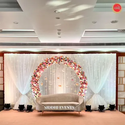 A perfect circle of pastel roses and hydrangeas frames a chic beige sofa, creating a stunningly romantic and elegant stage for a ring ceremony. The fairy light curtain adds a touch of sparkle to the backdrop.