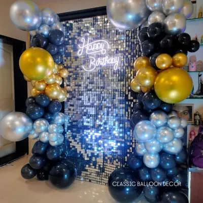 A stunning silver shimmer wall framed by black, gold, and silver balloon pillars. The 'Happy Birthday' neon sign adds a modern, celebratory glow.