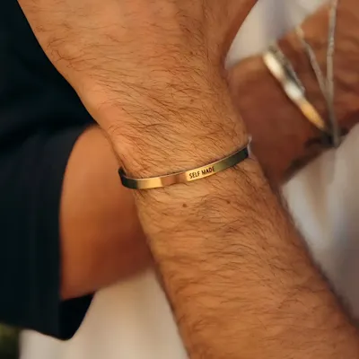 A man wearing a gold-plated cuff bracelet engraved with the powerful affirmation "SELF MADE". It's a bold statement piece for anyone proud of their journey.