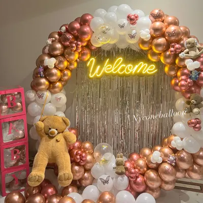 This welcome baby decoration features a beautiful ring arch made of rose gold and white balloons. A 'Welcome' neon sign, a shimmering backdrop, and teddy bear details make it a perfect surprise.