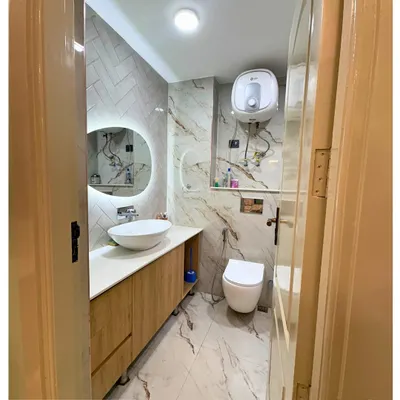 A clean and modern bathroom design featuring marble-finish tiles on the floor and walls. The floating wooden vanity, vessel sink, and backlit mirror create a spa-like feel.
