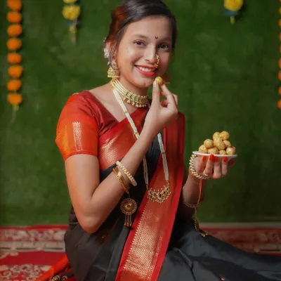 A classic Makar Sankrant portrait. The model is dressed in a traditional black and red saree, holding a plate of 'tilgul ladoo' against a festive backdrop of marigold flowers.