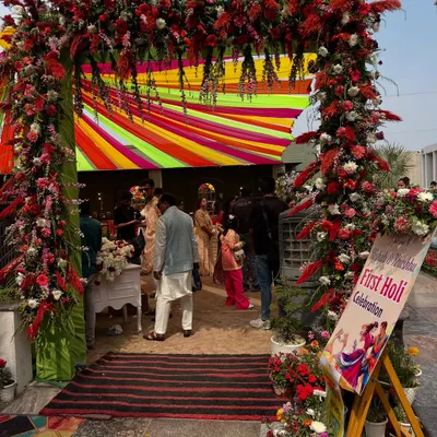 The perfect welcome for a Holi party! This grand entrance arch is bursting with red flowers and colorful fabric drapes, setting a festive and high-energy mood for the 'First Holi Celebration'.