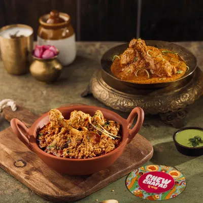 A rustic composition featuring Chicken Curry and Mutton Kheema for Chew Chapad. The use of traditional earthenware and brass bowls against a dark background enhances the rich, home-cooked feel of the dishes.