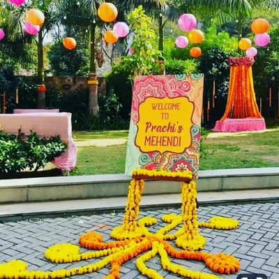 We created this vibrant outdoor Mehendi setup on a spacious lawn, designed to be both welcoming and picturesque. The personalized "Welcome to Prachi's Mehendi" sign, set on a wooden easel with marigold garlands, greets guests as they arrive. Overhead, colorful paper lanterns in pink and orange add a festive touch, complementing the decorated seating area visible in the background.