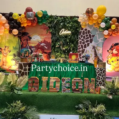 A grand Lion King safari theme for a first birthday. This elaborate setup features two distinct backdrops, artificial turf flooring, jungle foliage, and character cutouts of Simba, Timon, and Pumbaa.