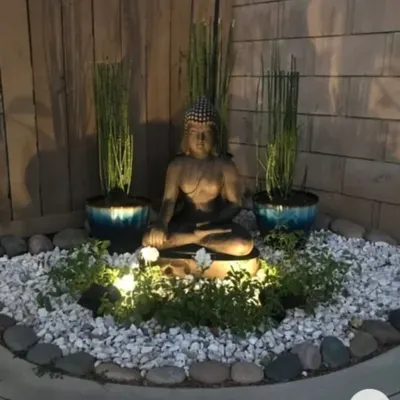 A serene Zen corner designed for evening relaxation. This setup features a meditating Buddha statue as the focal point, surrounded by white pebbles, uplighting, and minimalist plants for a tranquil ambiance.