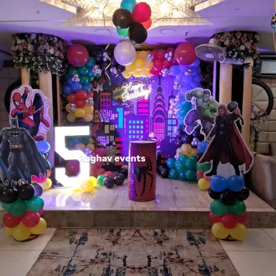 A full view of a superhero themed birthday party setup. This decoration features character cutouts of Thor, Batman, and Spiderman, a large marquee number '5', and a custom city backdrop with a neon 'Happy Birthday' sign.
