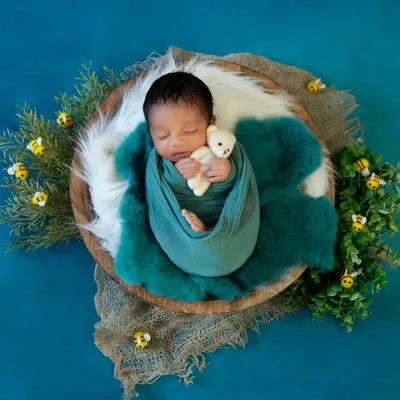 A top-down view of a baby boy swaddled in a teal wrap, holding a tiny bear. The setup includes greenery and small bee props against a rich blue backdrop, creating a nature-themed scene.