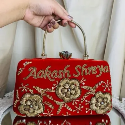 A vibrant red velvet clutch, personalized with the names 'Aakash' and 'Shreya' in beautiful golden script. The floral embroidery adds a touch of elegance to this custom bridal accessory.
