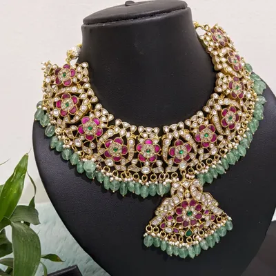 A breathtakingly beautiful Jadau necklace set with intricate floral motifs in pink and green, accented with uncut polki stones and natural strawberry beads. This is paired with matching chaandbaali earrings for a complete bridal look.