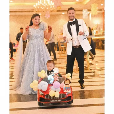 The birthday boy makes a memorable entrance in a toy car decorated with balloons, as his elegantly dressed parents dance alongside him. This shot captures the fun and festive atmosphere of the celebration.