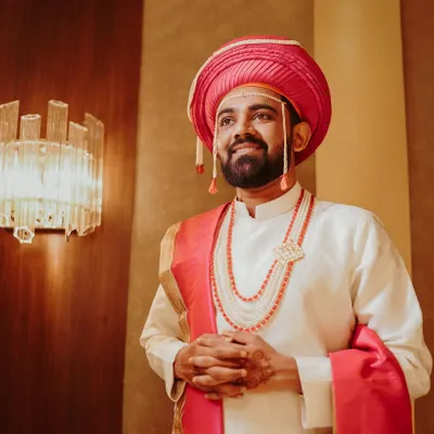 A classic portrait of a Marathi groom, looking dapper in his traditional wedding attire. The pink turban (pheta) and beaded jewelry add a touch of regal elegance.