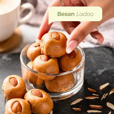 A hand reaches for a classic Besan Laddoo from a glass bowl. Made with slow-roasted besan and topped with an almond, it's the perfect comforting sweet to have with your evening chai.