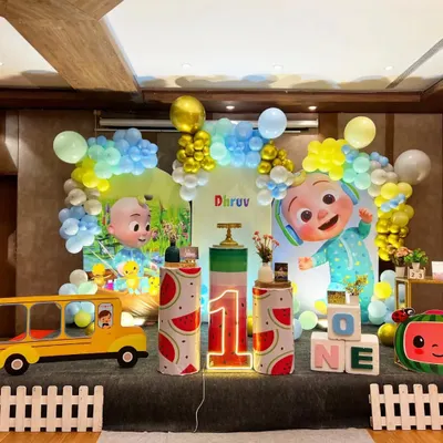 A vibrant Cocomelon themed first birthday party for Dhruv. This large stage setup includes multiple character cutouts, a watermelon-themed cake stand, a light-up number "1," and a yellow school bus prop.